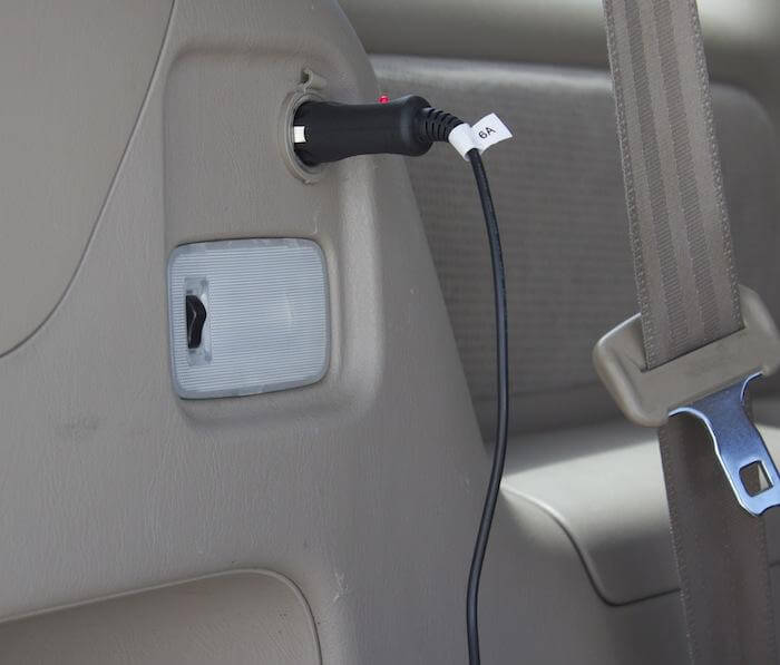 A Naxa 12 Volt DC Cord to power TVs with a universal connector is plugged into a car's cigarette lighter socket. The black cord has a "6A" label, with part of the seatbelt and car interior visible in the image.