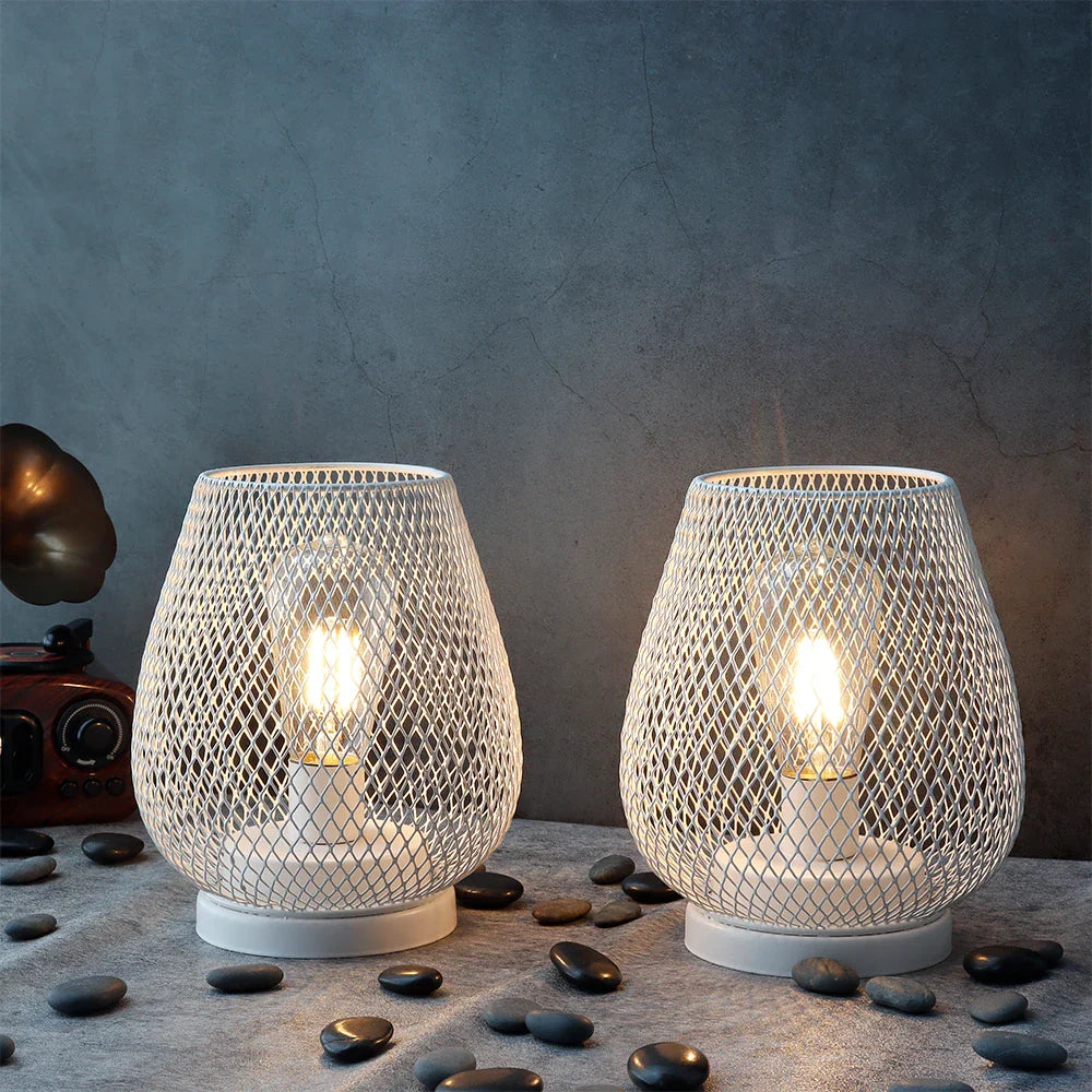 Two 6.7" tall battery powered outdoor table lamps from JHY DESIGN, featuring white mesh and warm bulbs, sit on a stone-textured surface among smooth black pebbles, with a vintage camera and small gramophone in the background.