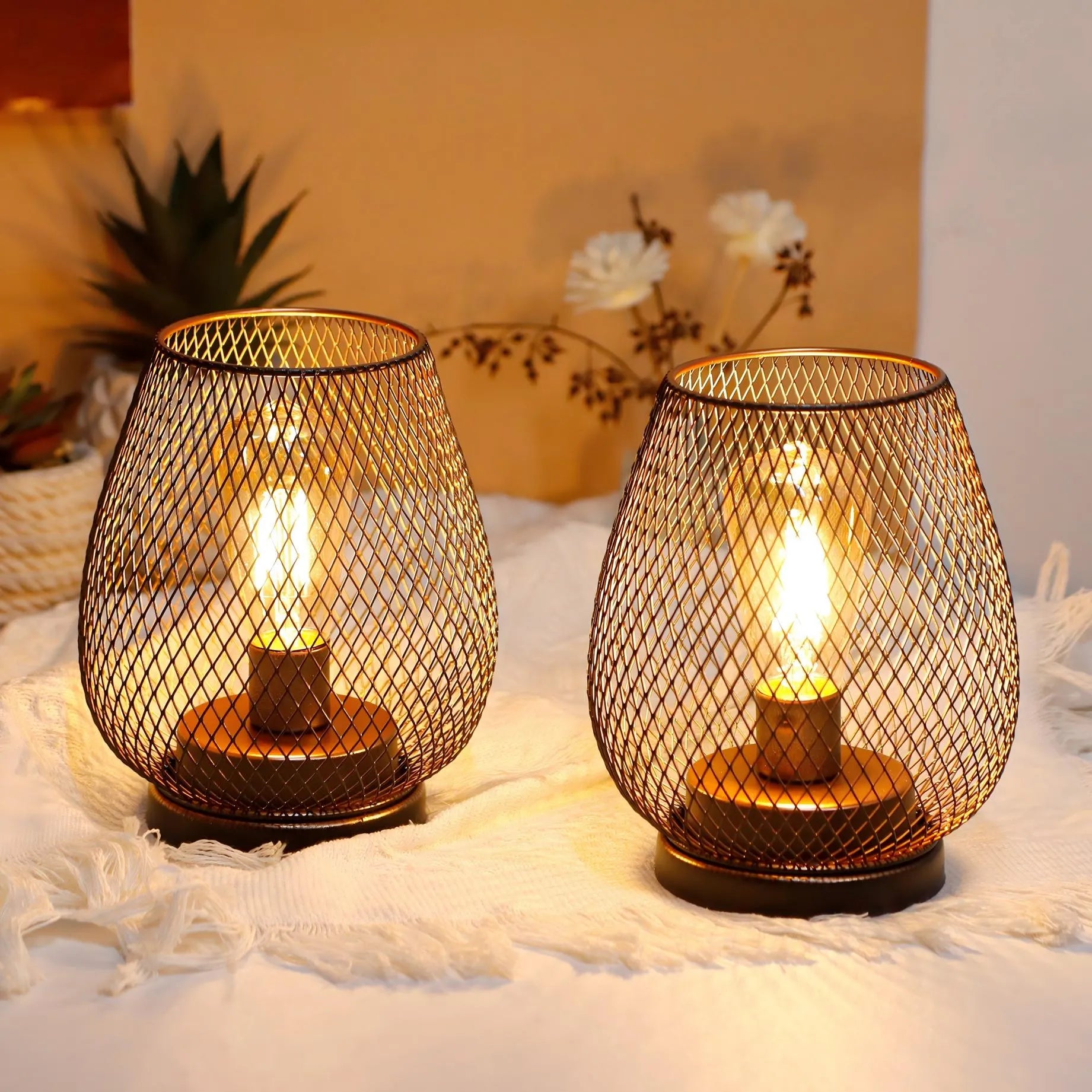 Two 6.7" tall battery powered outdoor table lamps from JHY DESIGN, featuring mesh wire covers and glowing bulbs, sit on a white textured cloth with blurred plants and flowers in the background for a cozy, warm ambiance.