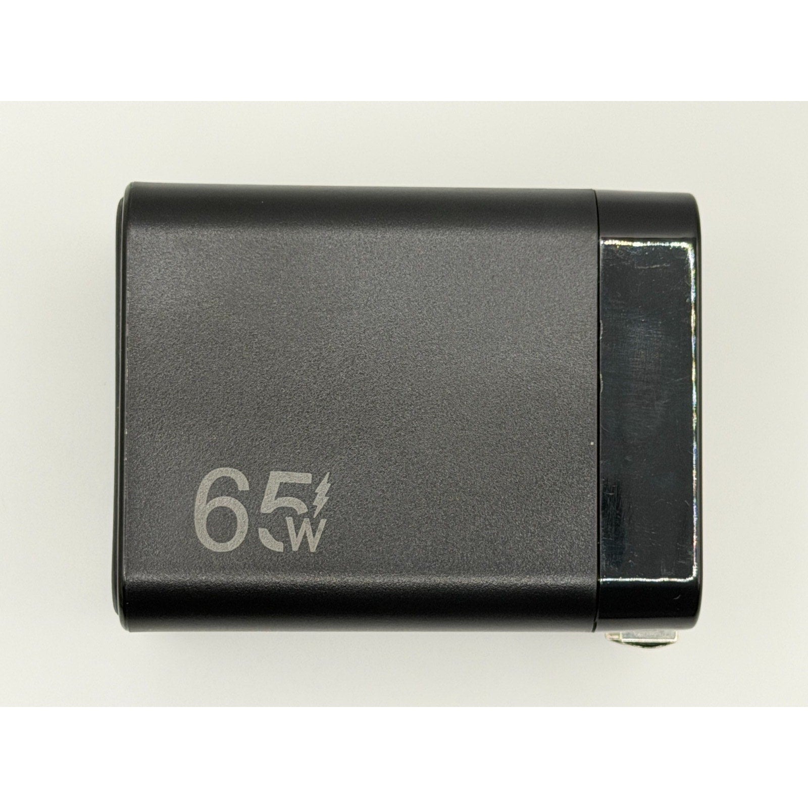 A 65W GaN USB-C Charger PD Fast Charge with foldable plug and built-in cable, black and rectangular, featuring a white lightning bolt symbol on the side; shown from above against a plain background.