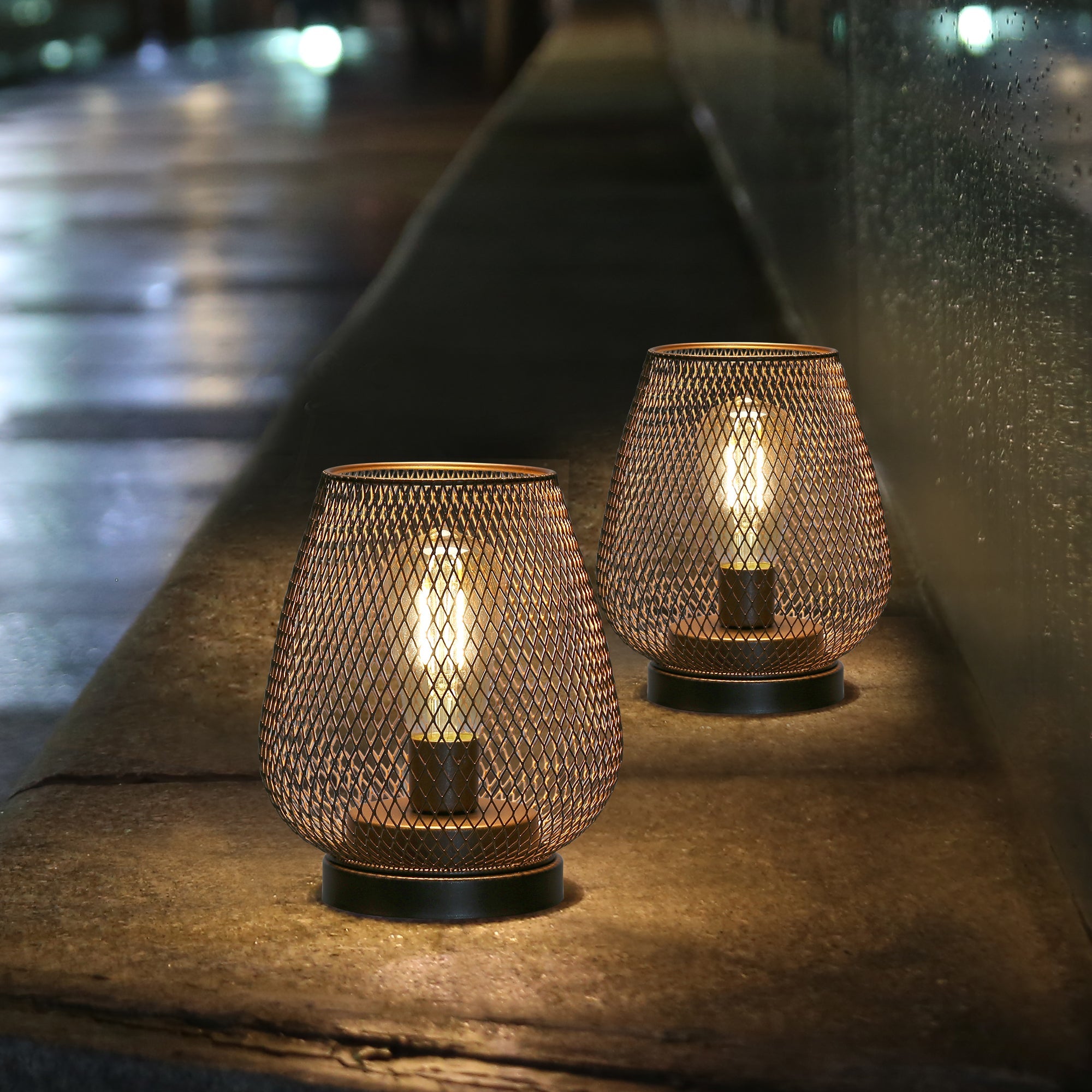 Two metal mesh lanterns, resembling the 6.7" Tall Battery Powered Outdoor Table Lamp, glow warmly on a stone ledge at night, their soft ambient light illuminating the blurred urban backdrop with inviting charm.