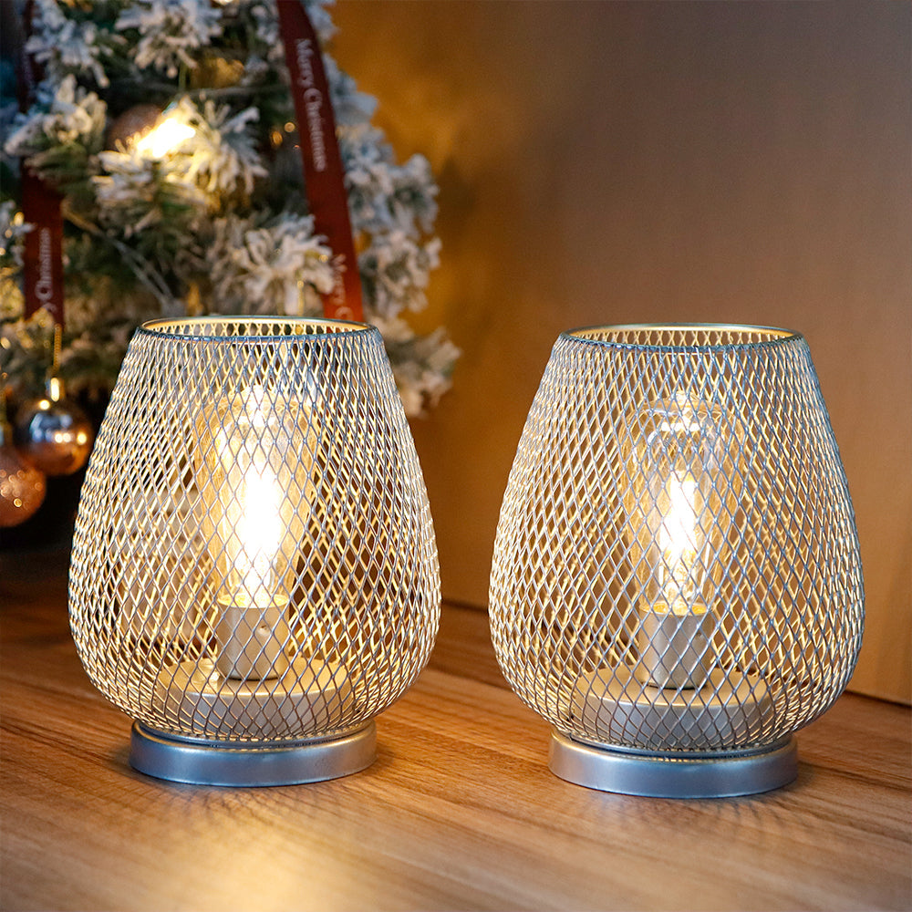 Two 6.7" tall battery powered outdoor table lamps by JHY DESIGN sit on a wooden surface, casting a warm glow. In the background, a Christmas tree with ornaments adds to the cozy, festive ambiance.