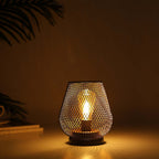 The 6.7" Tall Battery Powered Outdoor Table Lamp sits on a flat surface, its mesh metal shade softly illuminating a dim room, while faint shadows highlight part of a plant and books in the background.