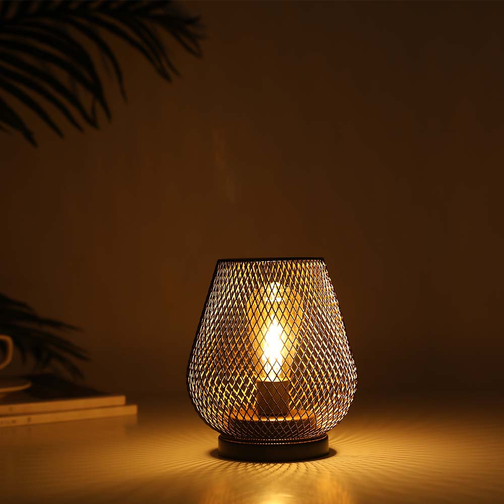 The 6.7" Tall Battery Powered Outdoor Table Lamp sits on a flat surface, its mesh metal shade softly illuminating a dim room, while faint shadows highlight part of a plant and books in the background.