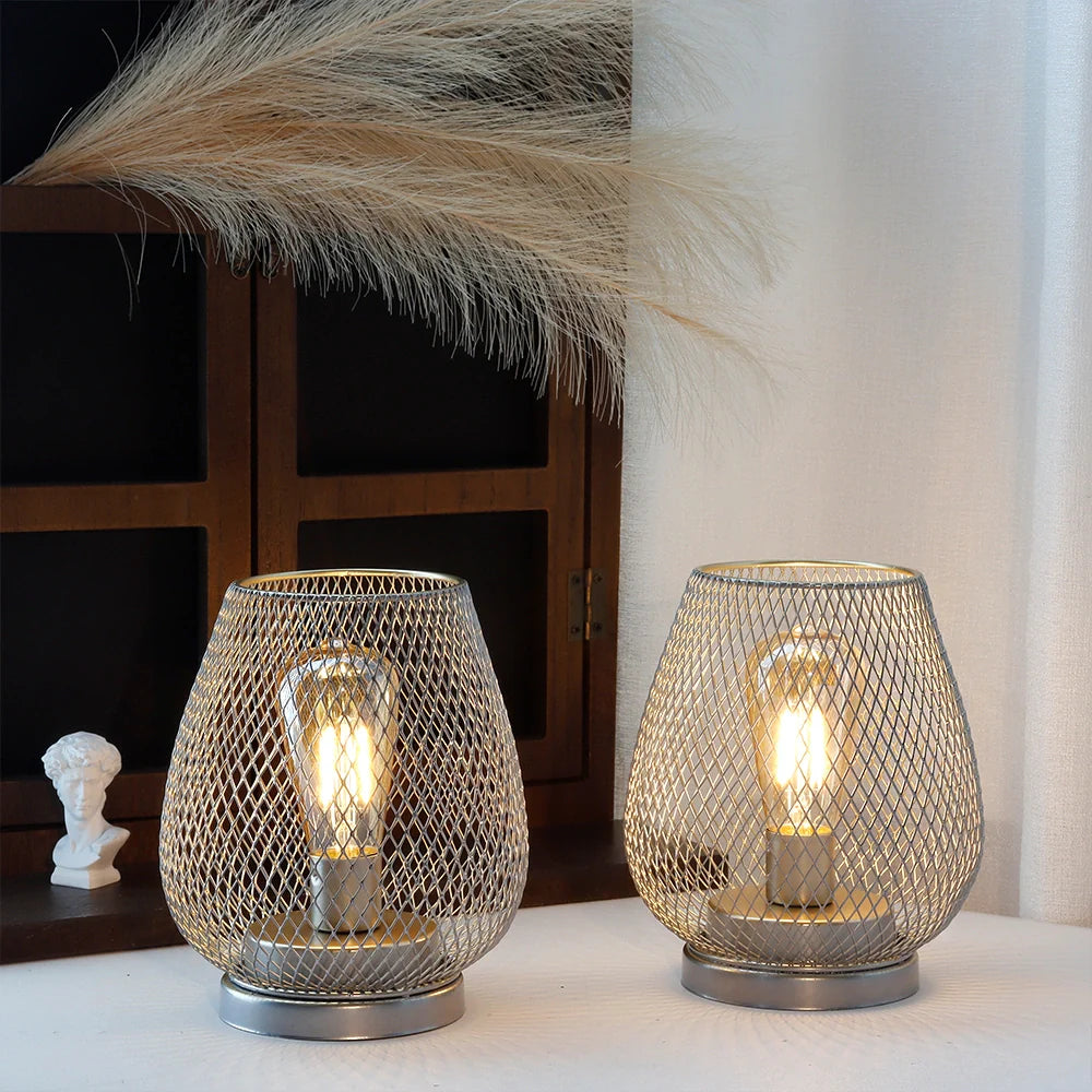 Two 6.7" tall battery powered outdoor table lamps with mesh metal designs and exposed bulbs emit a warm glow. Behind them are a window, a small white bust, pampas grass, and on the right side, white curtains add decoration.