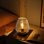 A 6.7" tall battery powered outdoor table lamp by JHY DESIGN with a mesh metal shade sits on a nightstand beside a bed with brown bedding. An open magazine lies beneath the cordless lamp, and part of a framed picture appears on the right.