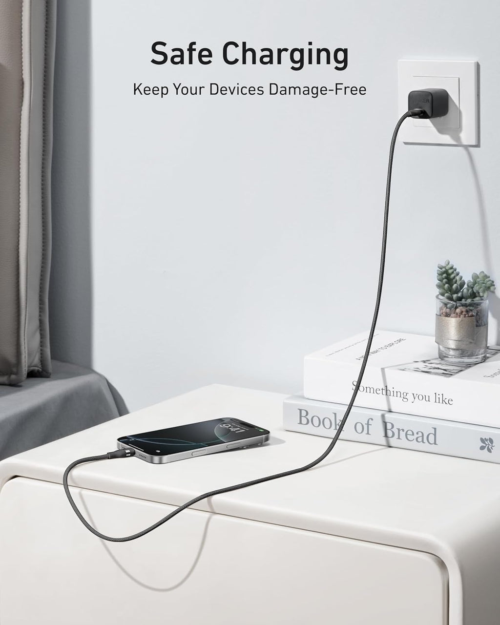A smartphone charges on a bedside table using the Anker USB-C to USB-C Cable (3 ft / 6 ft, 240W, Upcycled-Braided) plugged into a wall outlet. Two stacked books and a small potted plant sit beside it. Text above: "Safe Charging. Keep Devices Damage-Free.