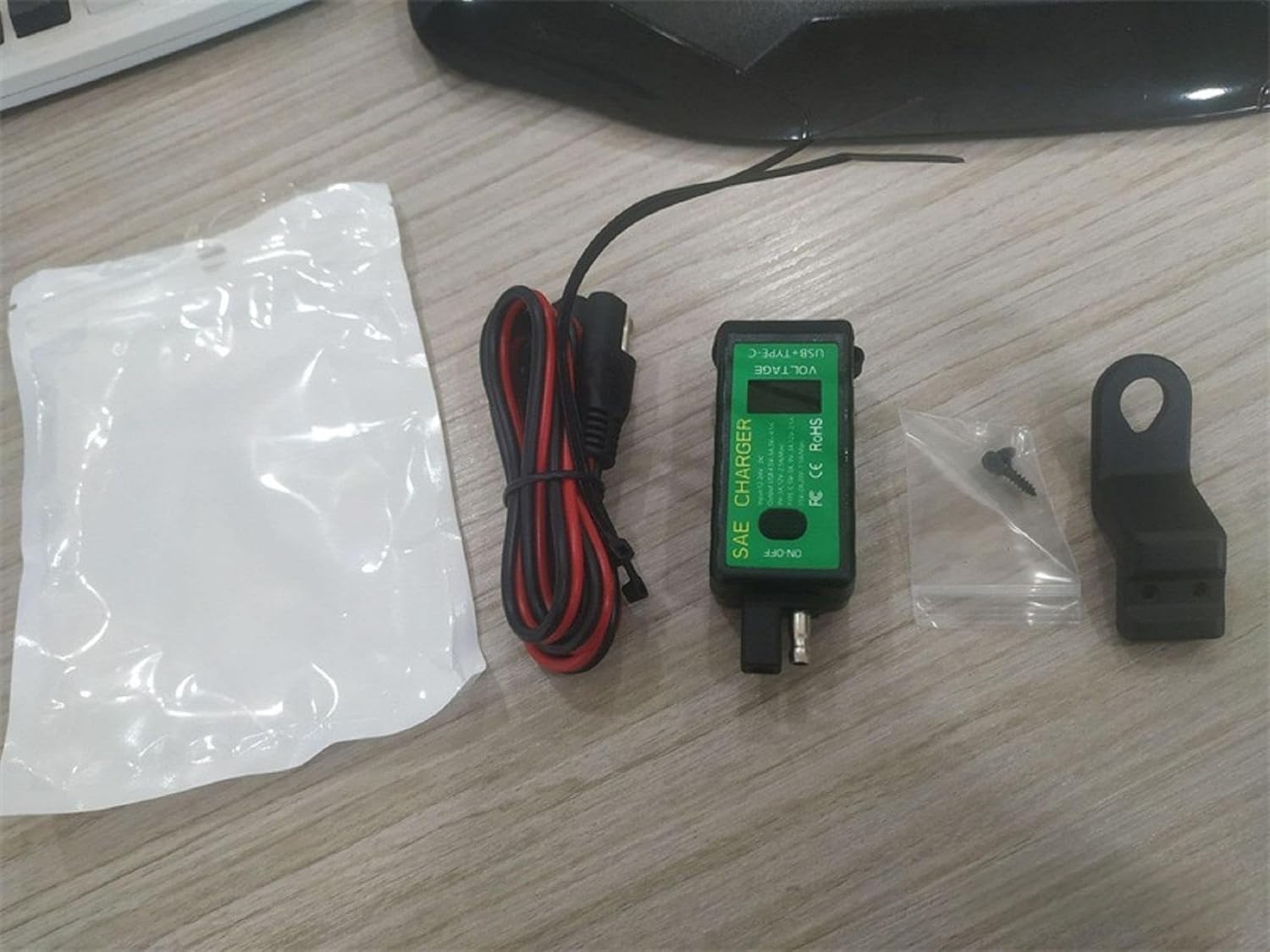 The Dual USB Motorcycle & Marine Charger – a 12V/24V waterproof USB power outlet with a built-in voltmeter and on/off switch – is displayed on a wooden surface, along with black and red cables, a small screw and wall anchor in a bag, a black mounting bracket, and white packaging.