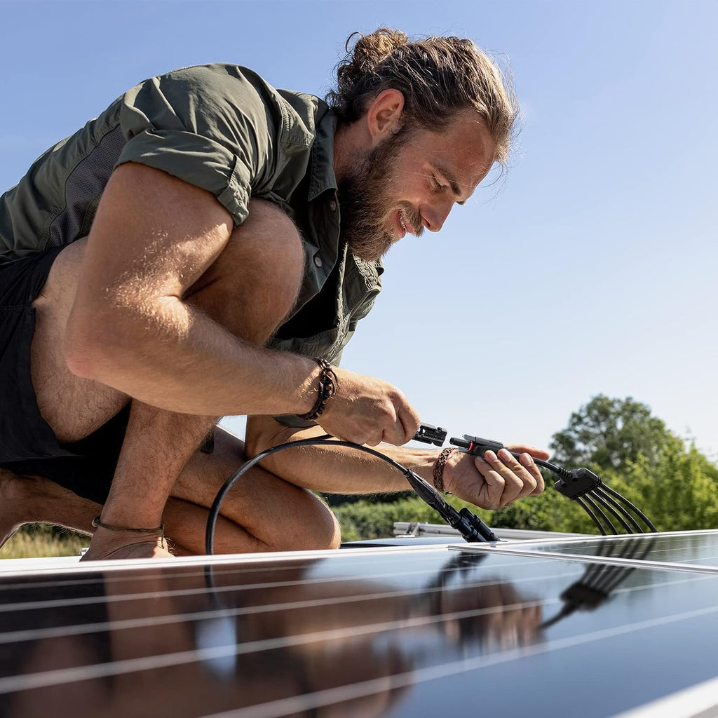 A man crouches outdoors, connecting wires with the BougeRV 1-to-5 Solar Y Branch Connector Cable Kit – MC4 Compatible Parallel PV Adapter (IP67 Waterproof, 30A 1000V) to a solar panel under a clear blue sky, with green trees in the background.
