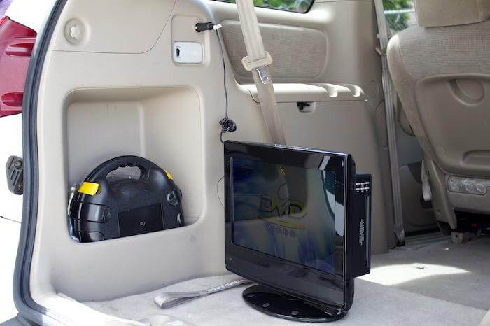 A Naxa 12 Volt DC Cord powers a portable DVD player in the back of a car via the cigarette lighter socket, with a car tool kit in a side compartment and a seatbelt hanging nearby.