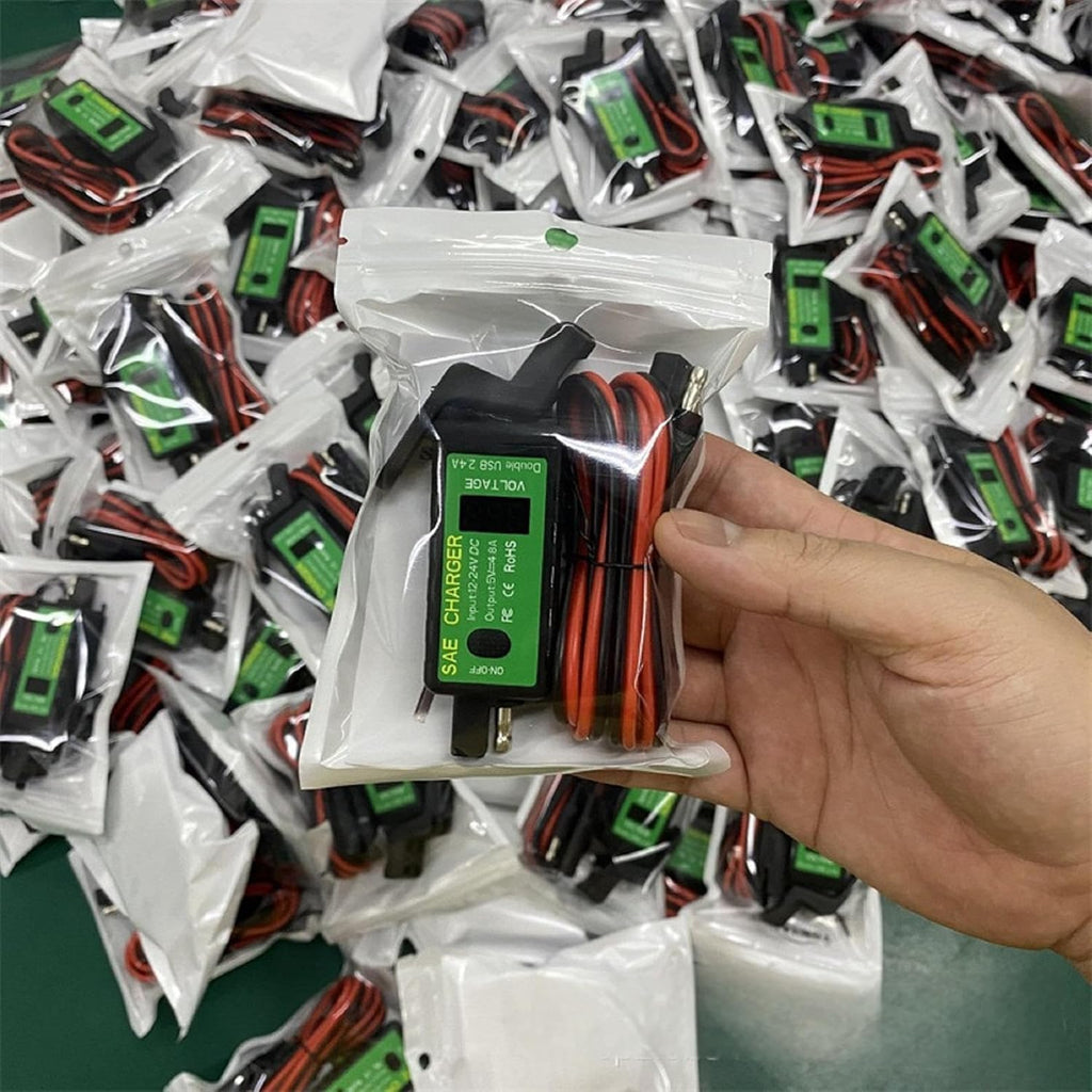 A hand holds a packaged Dual USB Motorcycle & Marine Charger – 12V/24V Waterproof USB Power Outlet with Voltmeter & On/Off Switch, featuring black and red cables. Numerous identical chargers in white pouches are scattered on a green surface in the background, making it ideal for anyone needing a reliable waterproof charging solution for their devices.