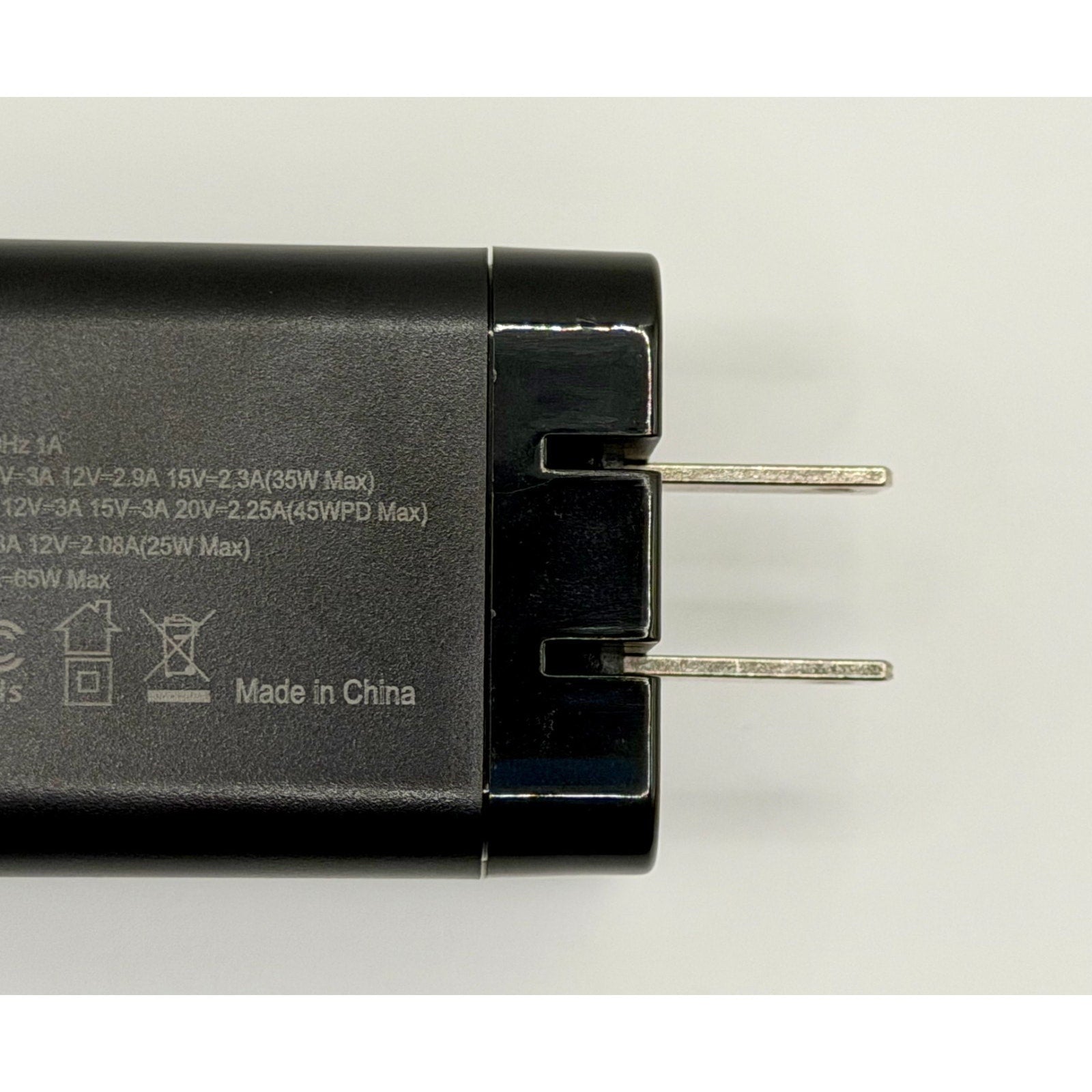 Close-up of a black 65W GaN USB-C Charger PD Fast Charge with foldable silver prongs, lying on a white surface. The brand new adapter shows white specs and "Made in China" printed on its side, plus a built-in cable.