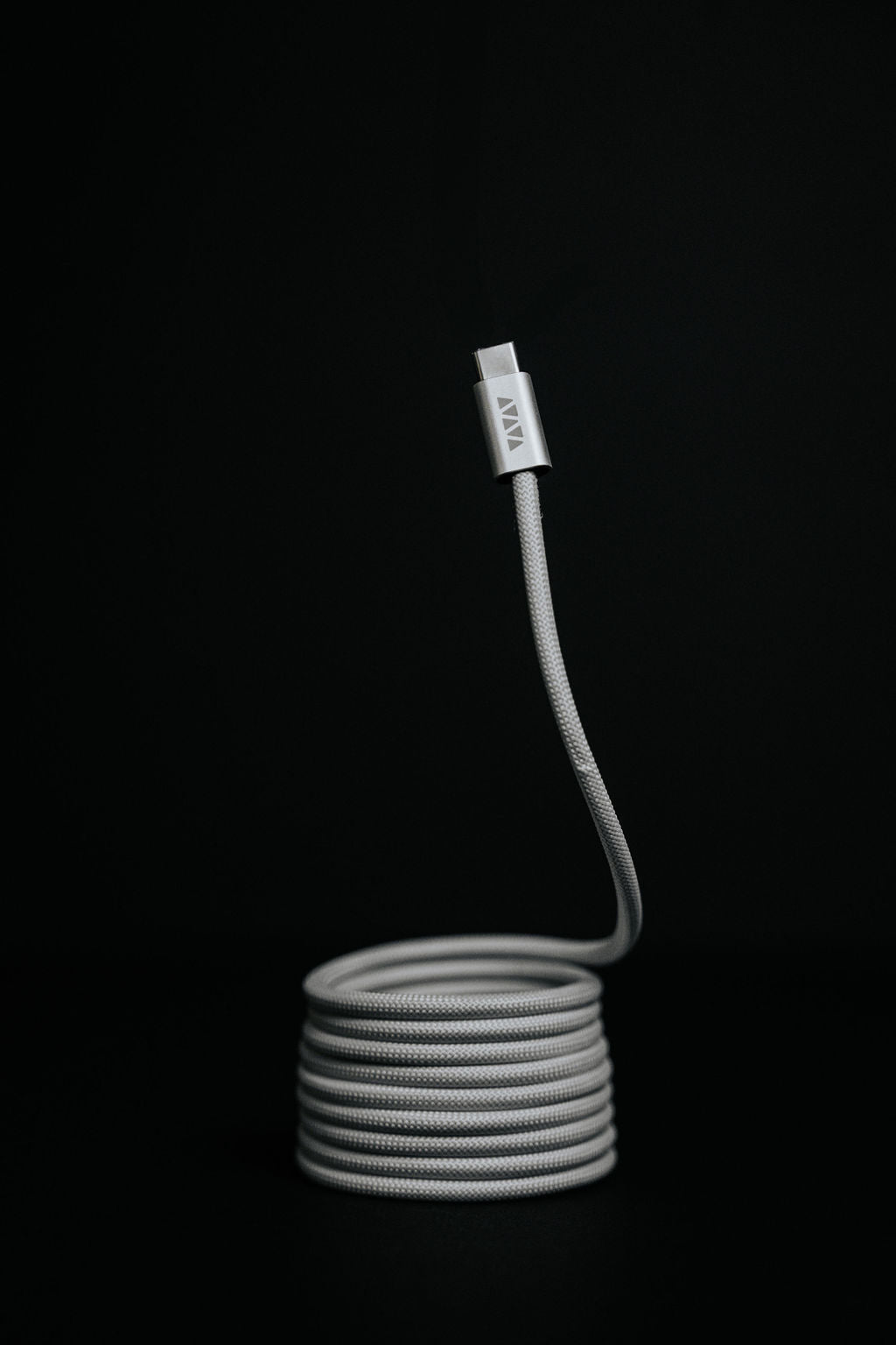 The Luxe Magnetic Cable, a coiled gray USB-C cable with a visible connector, stands upright against a black background—its heavy-duty build is perfect for fast charging.