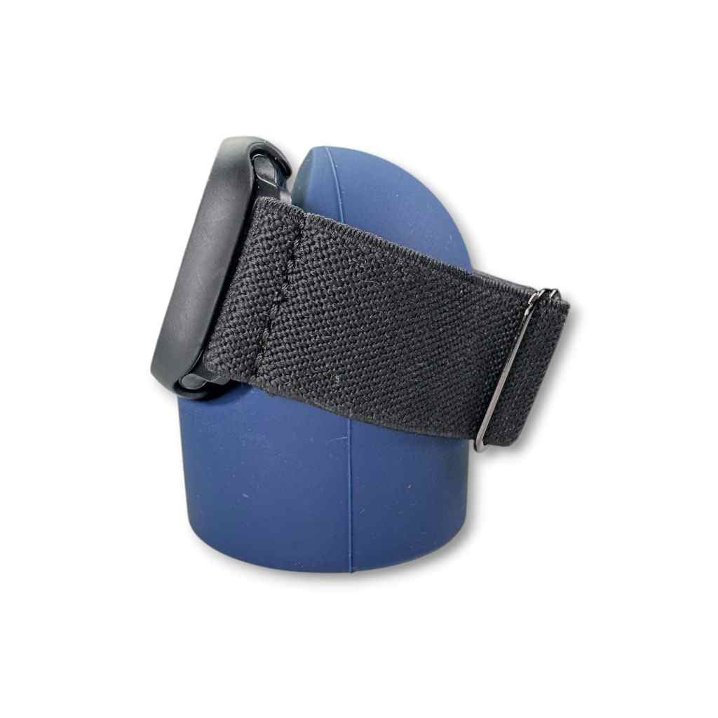 A black smartwatch with a fabric strap rests on a blue, rounded silicone Charging Stand against a white background. The watch face is turned away, showcasing the strap and side of the case.