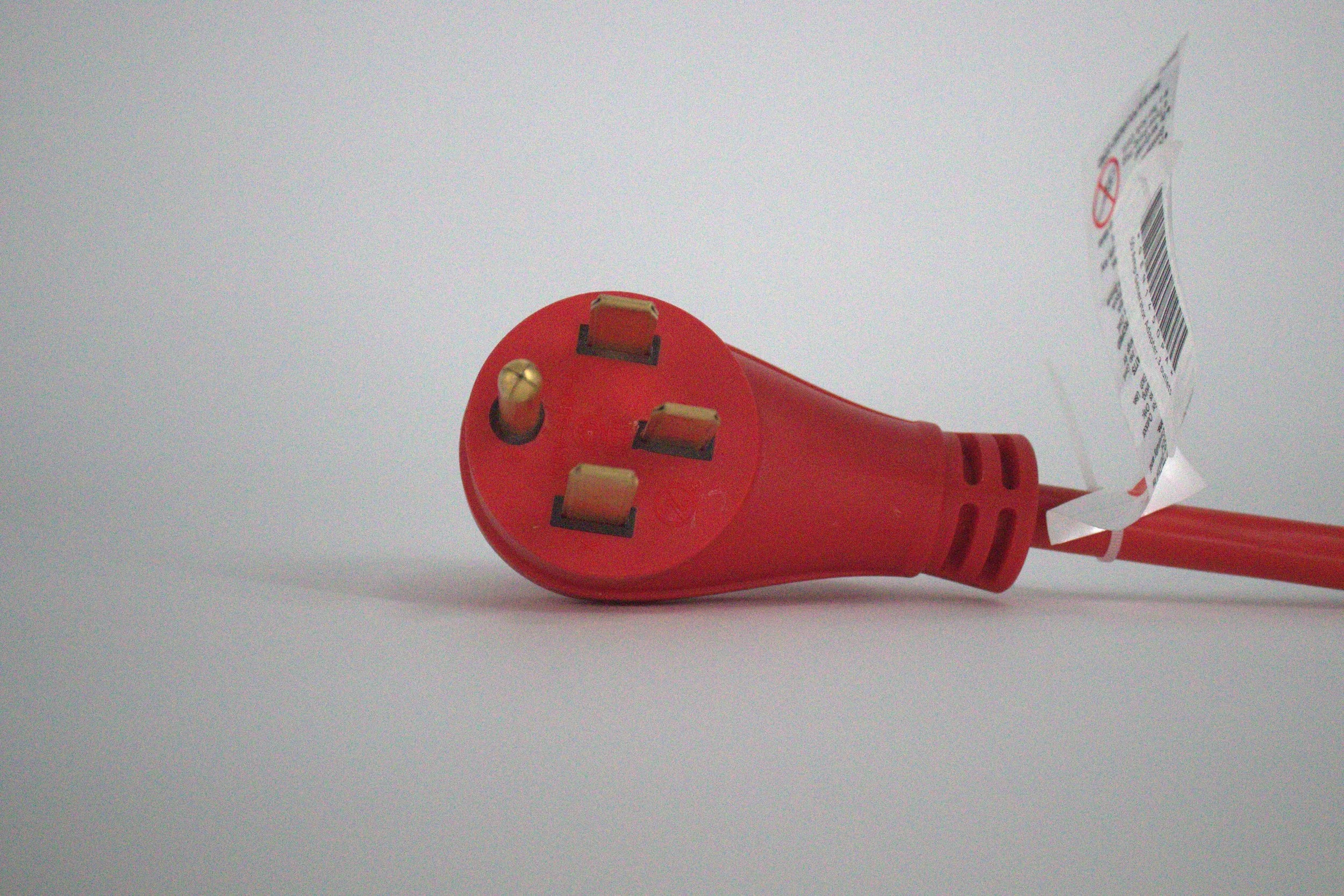 A close-up of a red 50 Amp Generator Adapter Cord 14-50P to 3x 5-15R Outlets with four brass prongs on a white surface. A tag with text is attached to the heavy-duty cord near the plug.