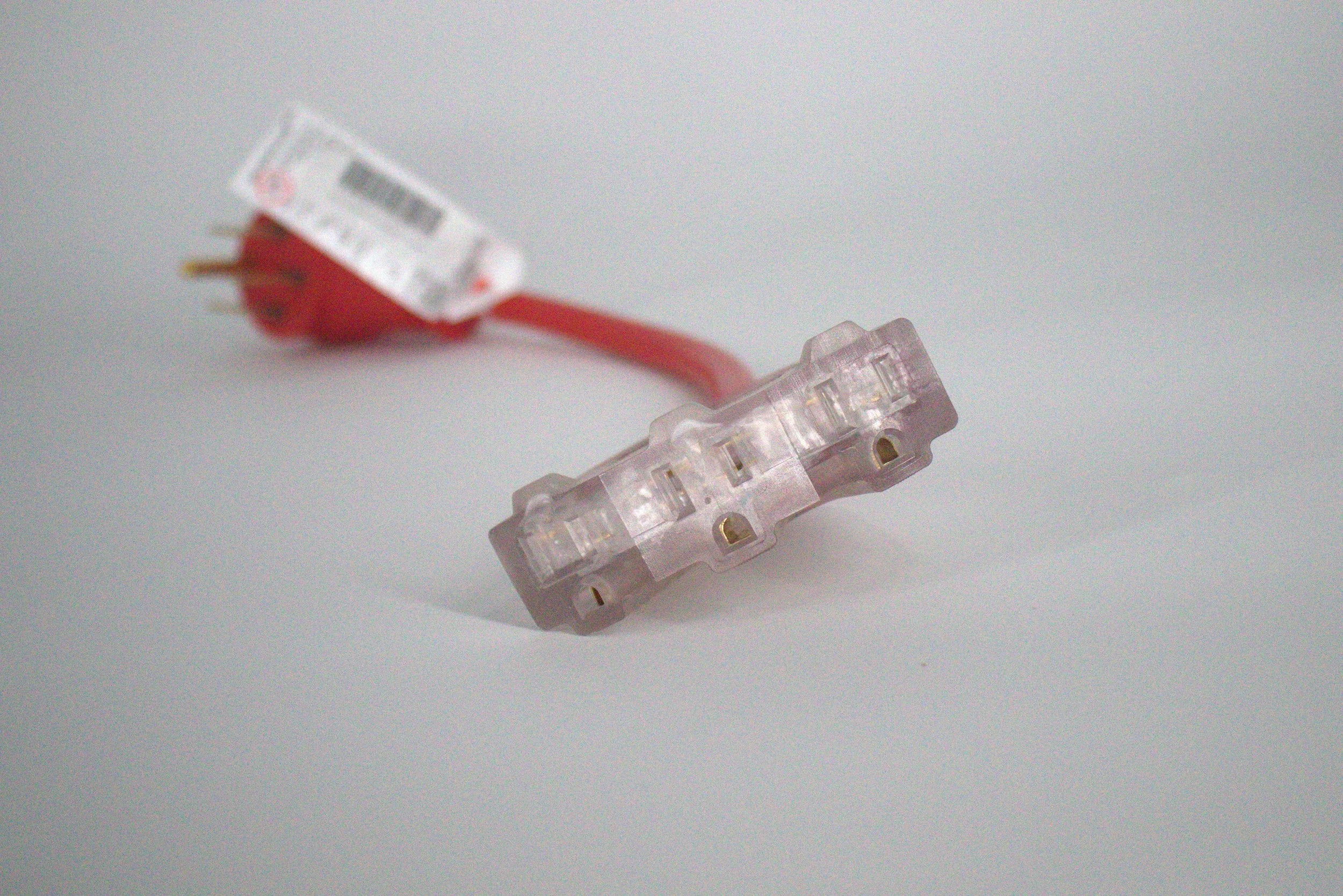A close-up of the 50 Amp Generator Adapter Cord 14-50P to 3x 5-15R Outlets, featuring clear three-outlet connectors and a red heavy-duty cord. The adapter and plug are visible but out of focus in the background.