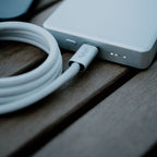 A close-up of a white portable power bank charging with the Luxe Magnetic Cable—a heavy-duty, braided USB-C cable—resting on wood. Four small LEDs on the power bank show its battery level.