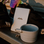 A Luxe Magnetic Cable with USB-C, plus a silver portable charger with a geometric logo, sits on a bench between black shoes in soft, warm light—ideal for fast charging on the go.