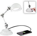 The Adjustable LED Study Lamp with Luminaire EyeCare USB Charging features white metal accents and delivers natural daylight illumination. A smartphone charges via a black cable connected to the lamp’s base, emphasizing its multifunctional design. An inset image shows a finger touching a button on the base, suggesting intuitive EyeCare lighting touch controls.