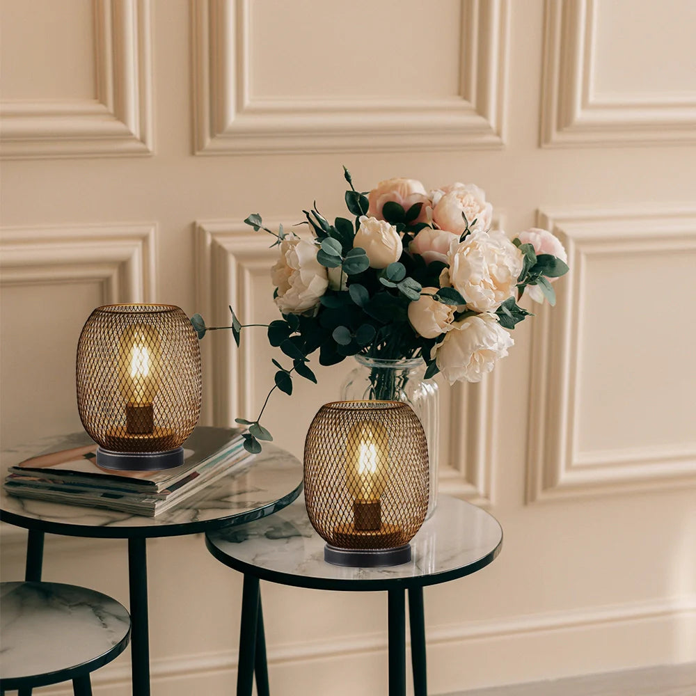 Two round side tables with marble tops display a set of 2 tall battery powered outdoor table lamps, mesh lantern-style, and stacked books. A vase with white and blush flowers adds elegance against cream-colored paneled walls.