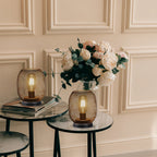 Two round side tables with marble tops display a set of 2 tall battery powered outdoor table lamps, mesh lantern-style, and stacked books. A vase with white and blush flowers adds elegance against cream-colored paneled walls.