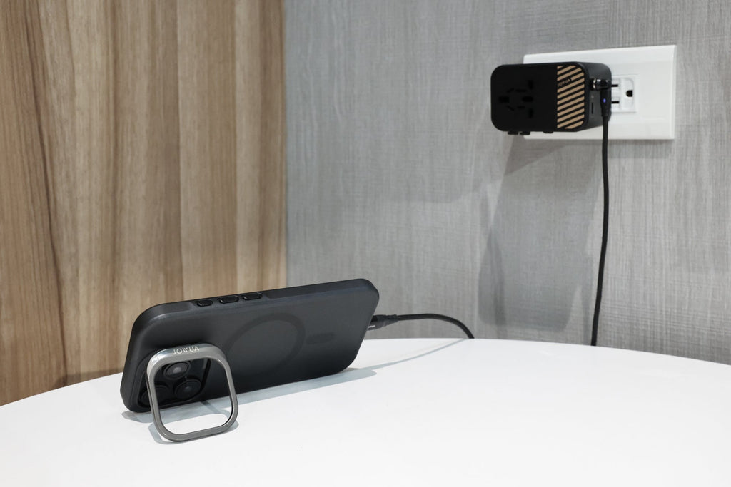 A smartphone charges on a white table, connected by cable to a 65W GaN Travel Adapter plugged into a wall outlet near a wooden and gray textured wall.