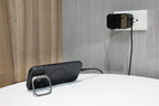 A smartphone charges on a white table, connected by cable to a 65W GaN Travel Adapter plugged into a wall outlet near a wooden and gray textured wall.