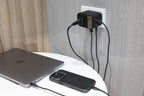 A 65W GaN Travel Adapter is plugged into a wall outlet, charging a laptop and a smartphone in a black case on a white table, with light wood and gray walls in the background.