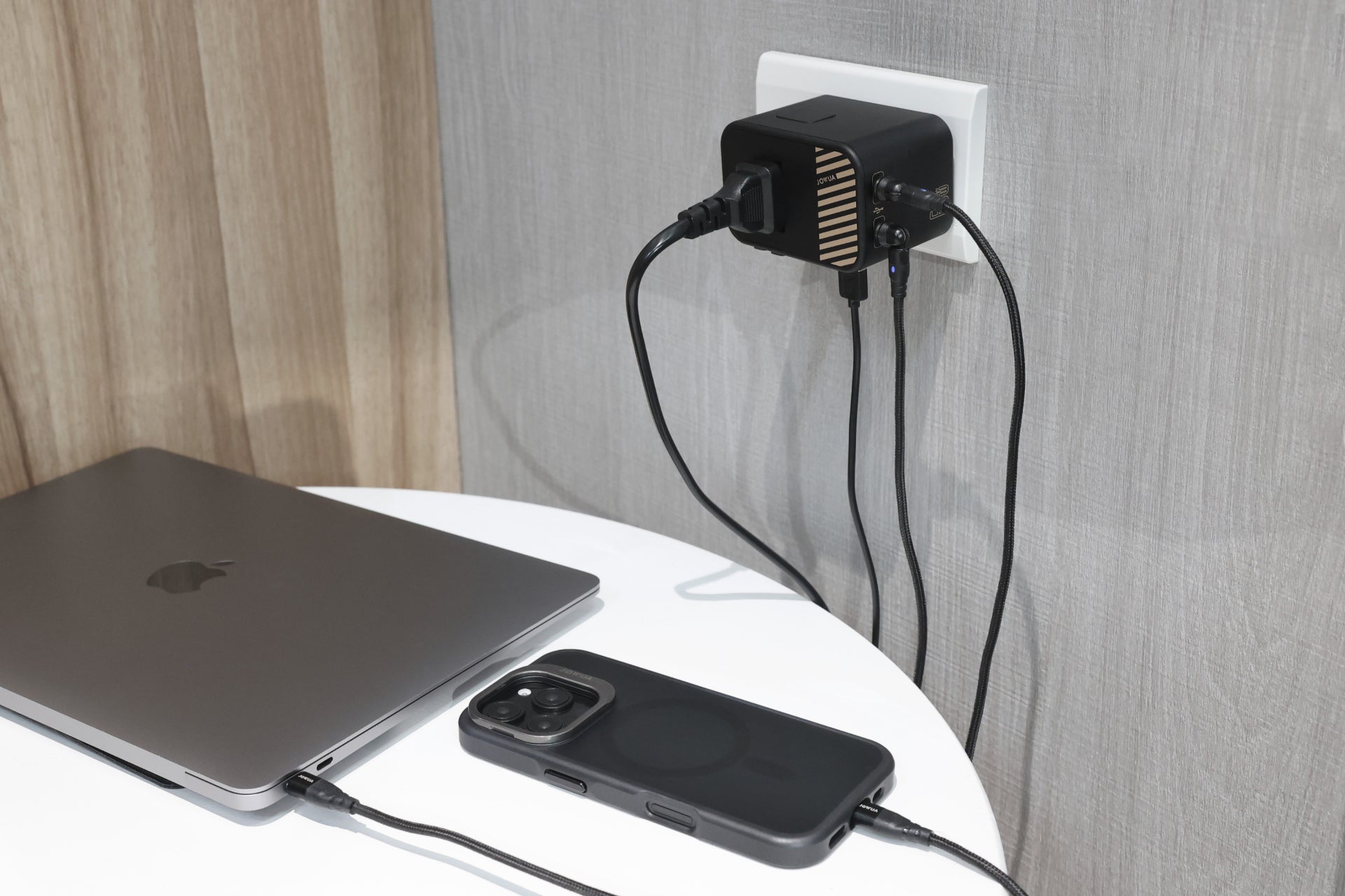 A 65W GaN Travel Adapter is plugged into a wall outlet, charging a laptop and a smartphone in a black case on a white table, with light wood and gray walls in the background.