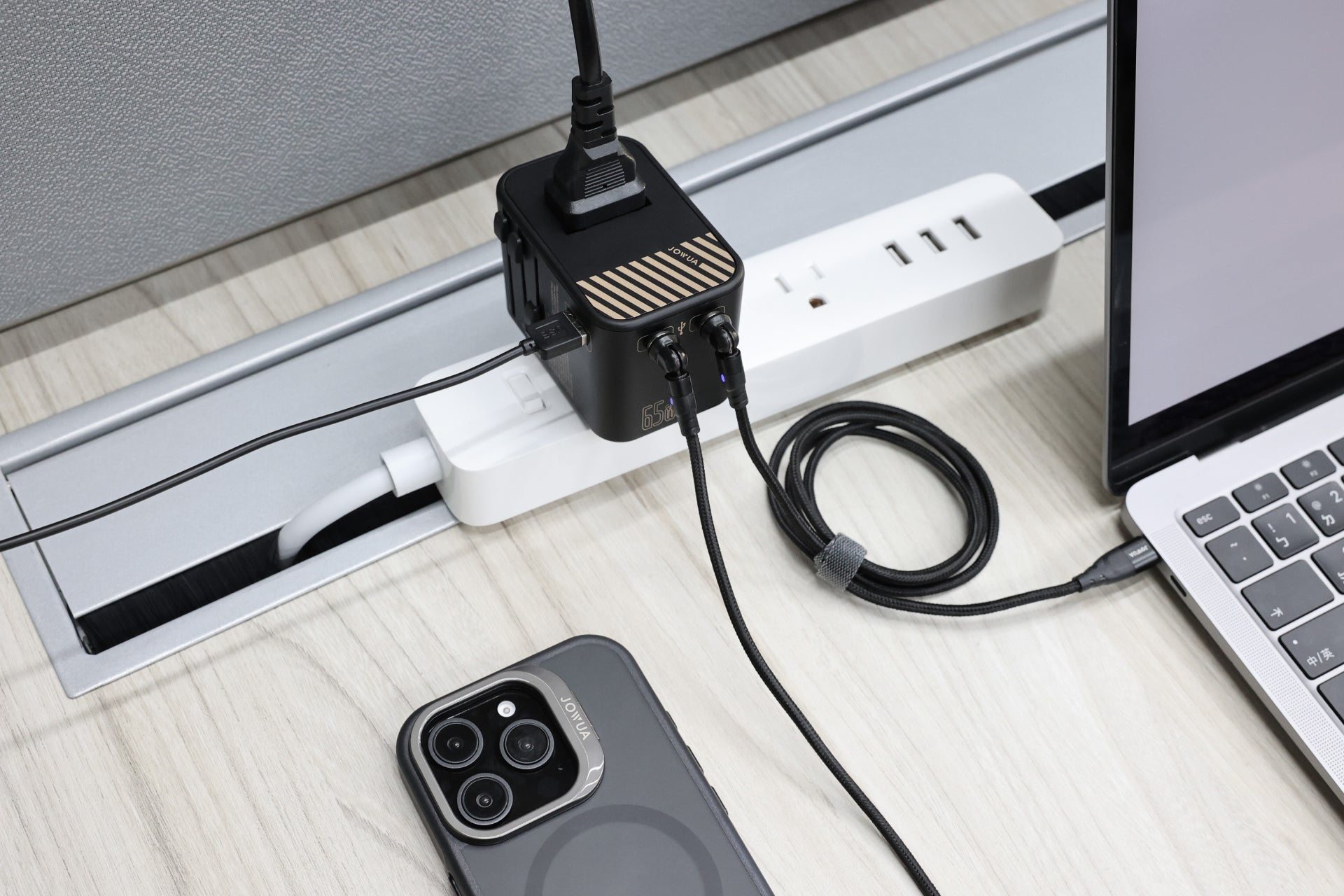 A modern, organized desk setup features a power strip with multiple devices plugged in, including the 65W GaN Travel Adapter, various chargers and cables, plus a laptop and smartphone placed nearby.